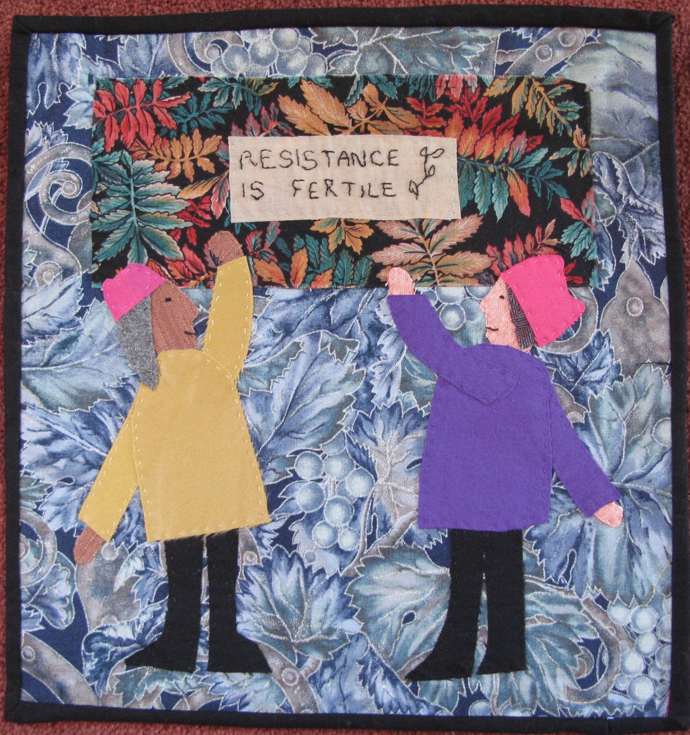 This fabric applique' piece shows to women wearing the symbolic pink hats of the Women's March. They are standing against a background of blue leaves and berries, holding up a banner bordered with more leave in fall colours. The words on the banner read:  Resistance is Fertile.
 