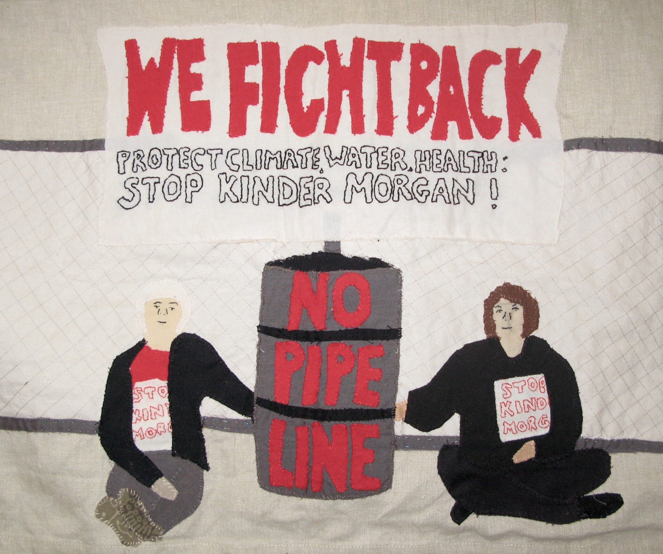 Two women are sitting on the ground in front of a chain link fence. They are both wearing shirts that say "Stop Kinder Morgan" and their hands are chained to the fence behind a barrel that says "No Pipe Line". Above them is a banner that reads: "We Fight Back. Protect Climate, Water, Health: Stop Kinder Morgan !"